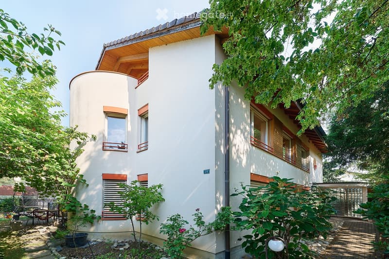 Two-story house with white walls, wooden roof, and multiple windows. The house is surrounded by greenery, including trees and shrubs, with a small patio area featuring outdoor furniture.
