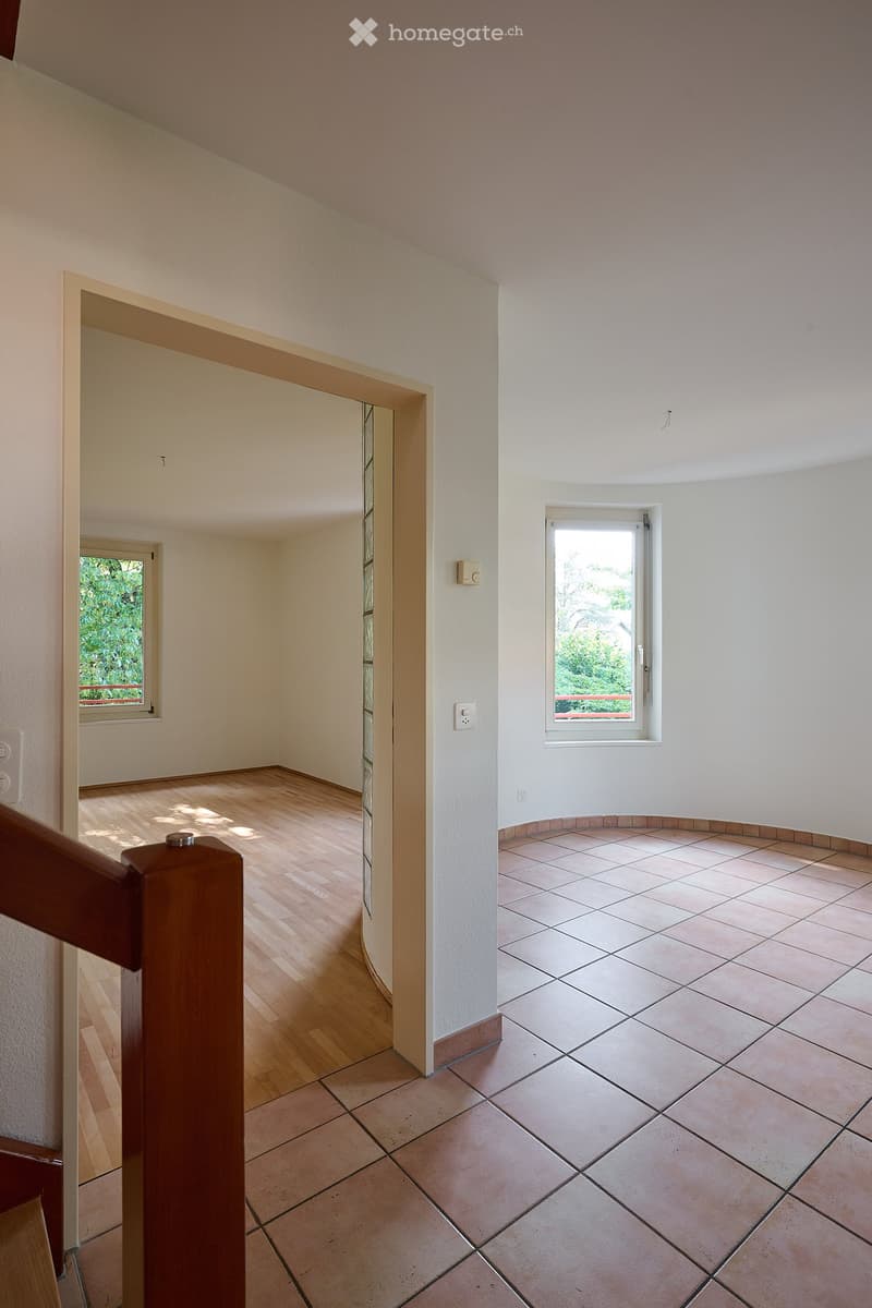 Empty hallway with wooden flooring and tiled section, white walls, and two windows letting in natural light.