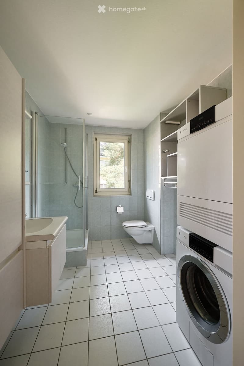 Modern bathroom with white tiled floor and walls, featuring a sink, toilet, shower, and stacked washing machine and dishwasher unit.