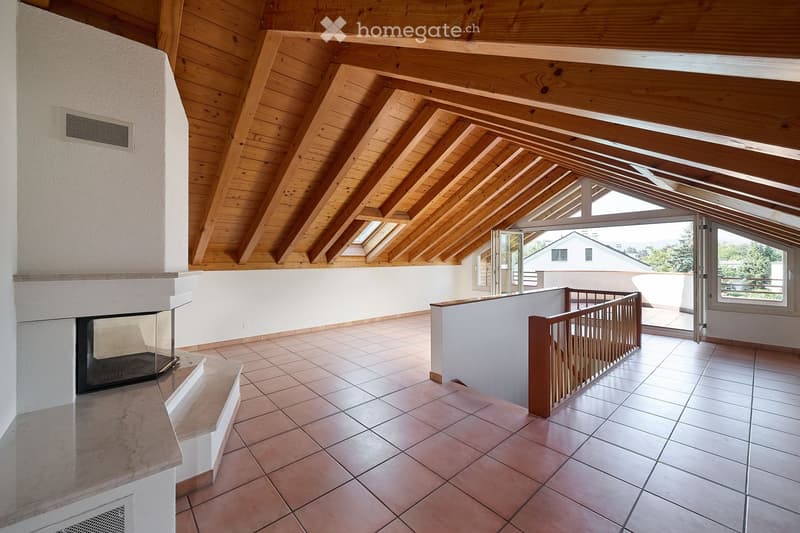 Bright hallway with wooden slanted ceiling, tiled floor, fireplace on left, open doorway leading to outdoor area, and wooden railing.
