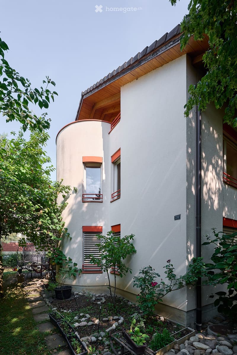 A modern, white house with a curved facade, surrounded by a well-maintained garden with potted plants, small trees, and a seating area with chairs and a table.