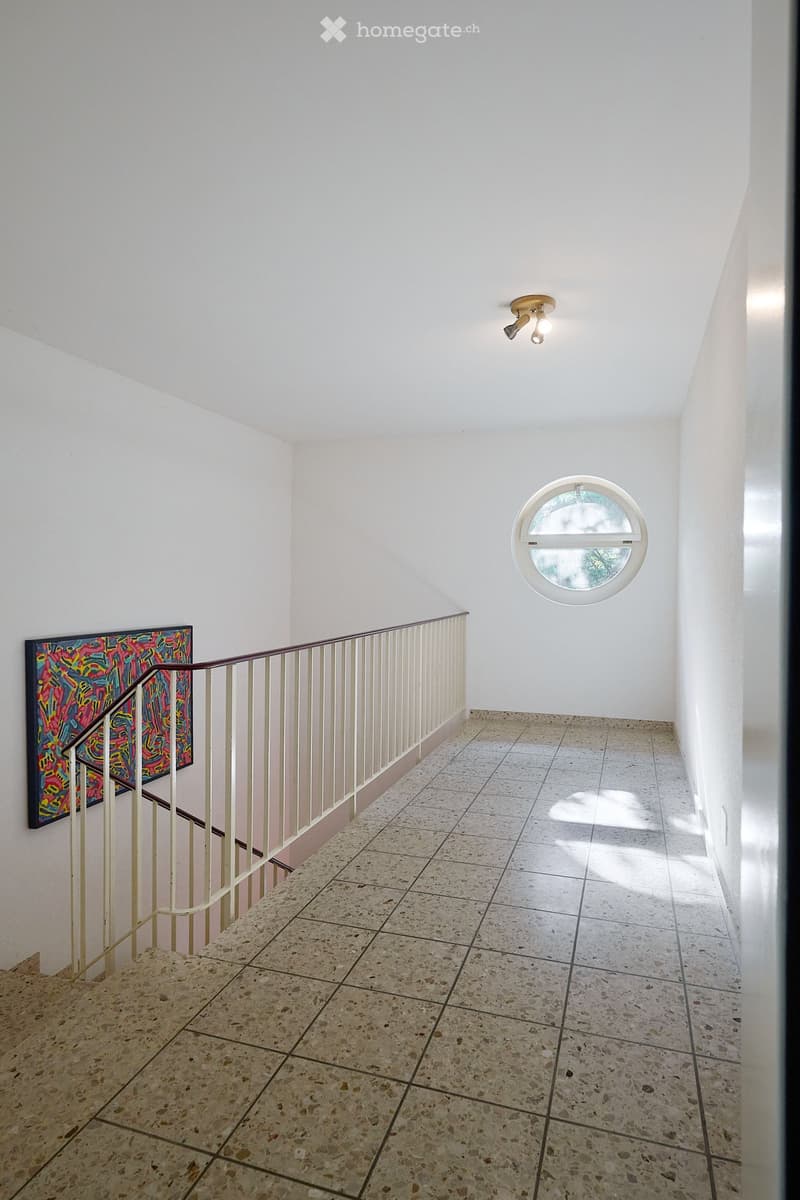 Staircase with beige railing, colorful artwork on wall, round window, and modern ceiling light fixture.