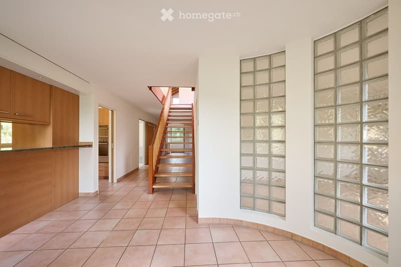 Bright hallway with tiled flooring, wooden staircase leading upwards, glass block walls, and wooden cabinetry on the left side.