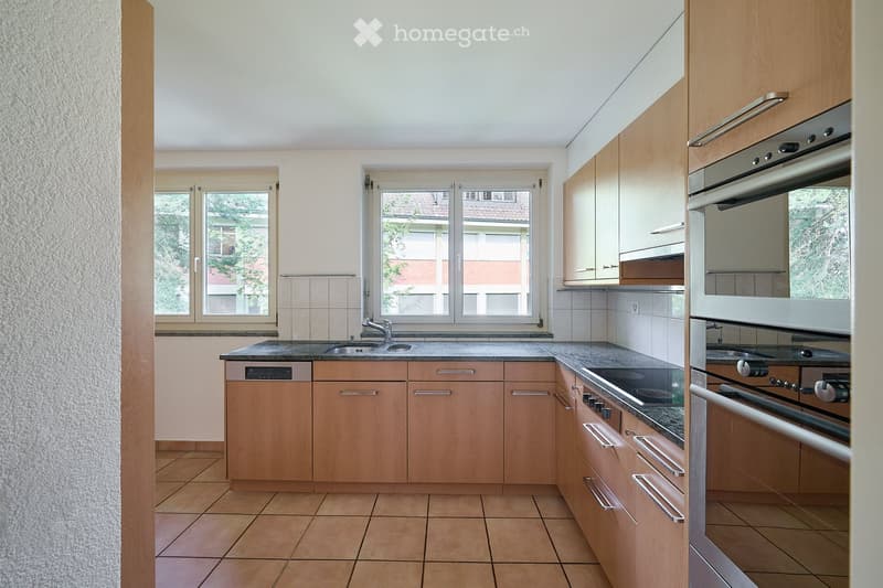 Modern kitchen with light wooden cabinets, granite countertops, stainless steel appliances, and large windows.