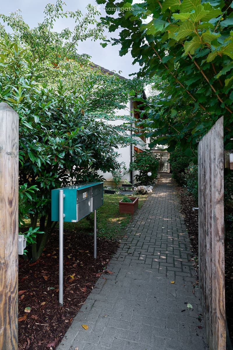 A narrow garden pathway lined with lush greenery, leading to a house entrance. The pathway is paved with bricks and bordered by wooden fences. There is a mailbox on the left side and a small planter in the middle of the path.