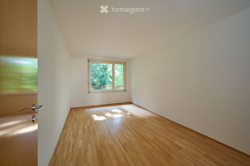 Empty room with wooden flooring, white walls, and a window with a view of greenery outside.