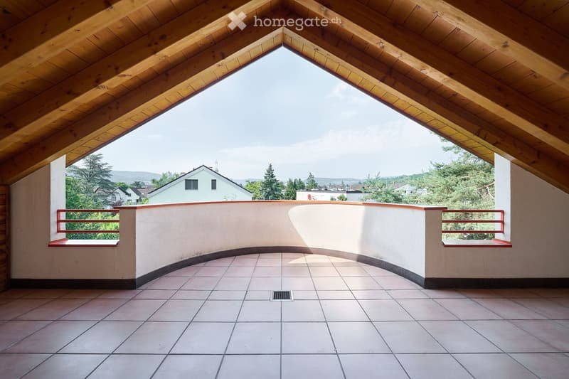 Balcony with wooden slanted ceiling, tiled floor, and a view of the surrounding area with trees and houses.