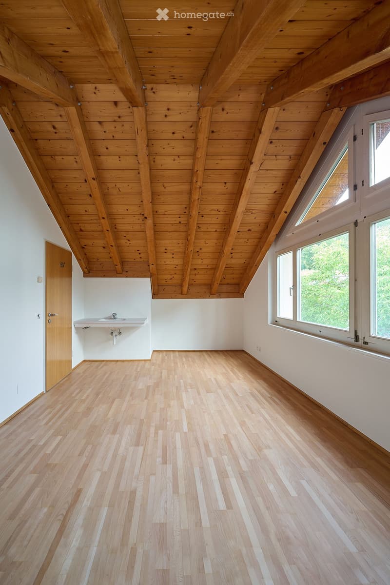 Bright, empty room with wooden floors and exposed wooden beams on the ceiling. Features large windows with white frames and a small sink area with a faucet.