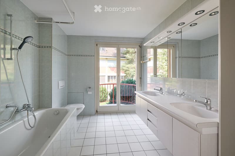 Modern bathroom with a bathtub, toilet, double sink, large mirror, and a showerhead. The room features tiled walls and floor, and a sliding glass door leads to a balcony.