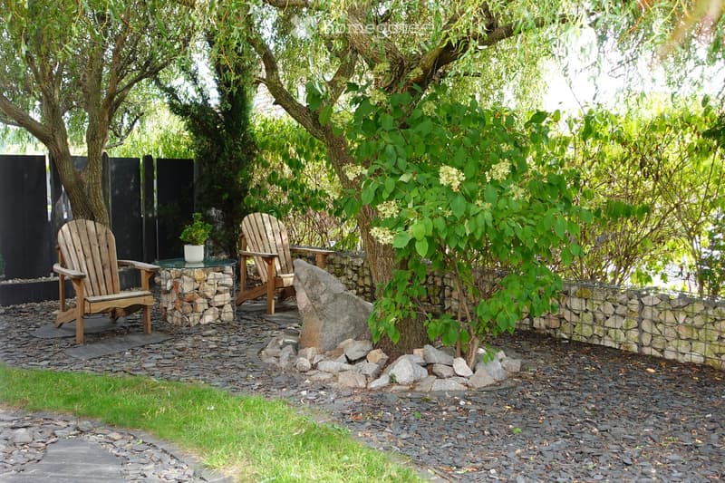 A serene garden area with a stone pathway, two wooden chairs, a small round table with a potted plant, and a large tree providing shade. The area is enclosed by a stone wall and wire fence, with scattered rocks and greenery.