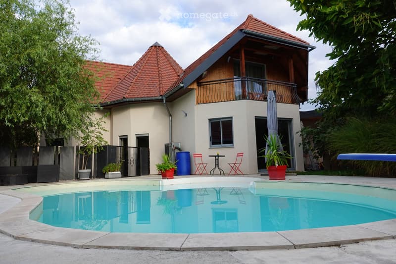 A modern house with a red-tiled roof and a balcony, surrounded by greenery. A swimming pool is visible in the foreground, with outdoor furniture including red chairs and a small table. Potted plants and a closed umbrella are near the house entrance.