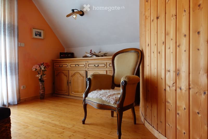 Cozy attic room with wooden floors, slanted ceiling, and warm lighting. Features a wooden cabinet, a plush armchair, a vase with flowers, and a picture frame on the wall.