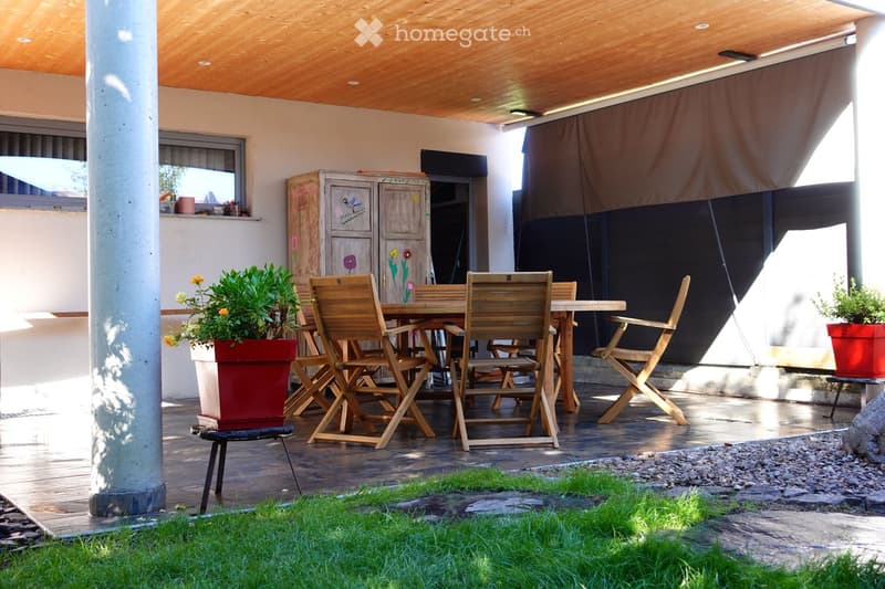 Wooden outdoor seating area with a table, chairs, and potted plants, featuring a covered wooden ceiling and a grassy area.