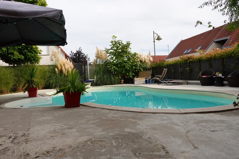 Outdoor area with a swimming pool, surrounded by potted plants, lounge chairs, and a covered patio area. The pool is rectangular with a smooth concrete deck, and there are lounge chairs and a small table near the pool.