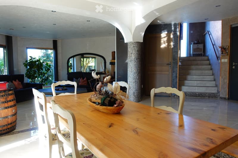 Bright, open-plan living and dining area with a long wooden dining table surrounded by white chairs, a sofa with cushions, a decorative bowl centerpiece, and a large mirror on the wall. The space features large windows and a staircase leading to an upper