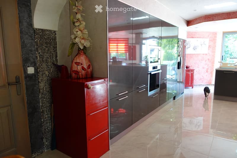 Modern kitchen with sleek, dark grey cabinets, a red sideboard, and a vase with flowers on top. The floor is tiled, and the kitchen features stainless steel appliances, including a refrigerator and an oven.