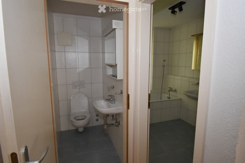 A small bathroom with white tiled walls and floor, featuring a toilet, sink, and bathtub. The space is compact with a door leading into the room.