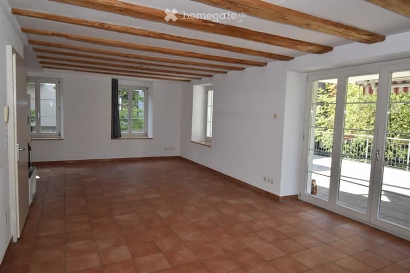Empty room with wooden ceiling beams, tiled floor, large windows, and sliding glass doors leading to a balcony.