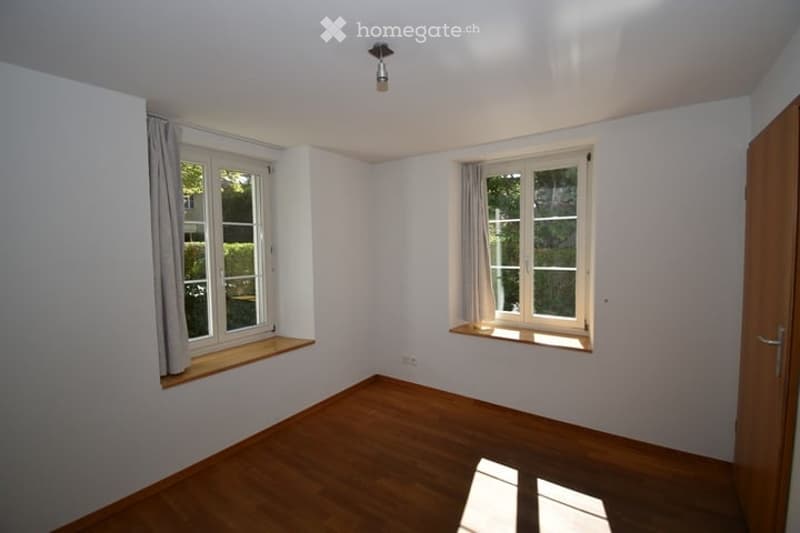Empty room with wooden flooring, white walls, and two windows with wooden sills and grey curtains.