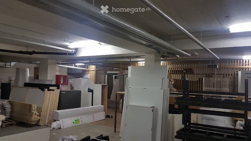 Empty room with exposed pipes and lighting, containing various construction materials and storage items, including wooden planks, cardboard boxes, and metal shelving units.