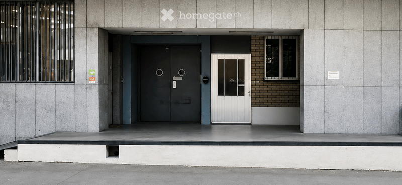 Modern building entrance with a raised ground floor, featuring a large grey door, a smaller white door, and a small window to the right.