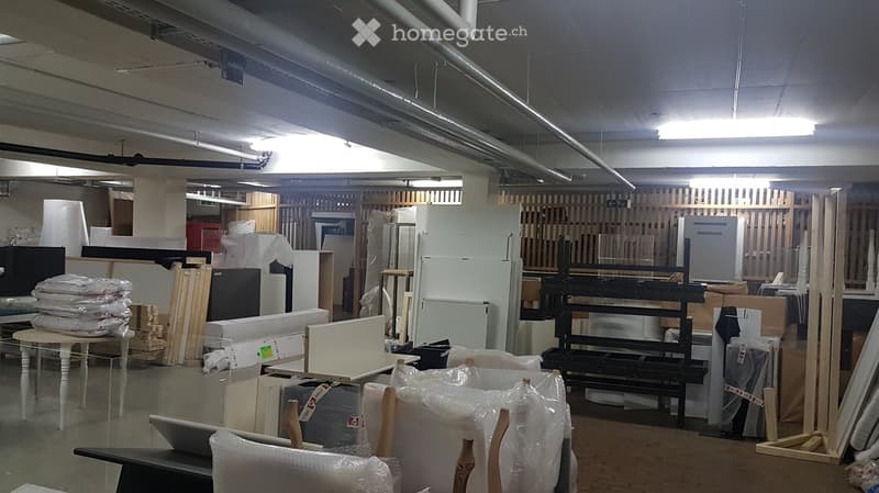 Empty warehouse or storage space with exposed pipes and lighting, containing various unpacked furniture and materials covered in plastic wrap and stacked against the walls.