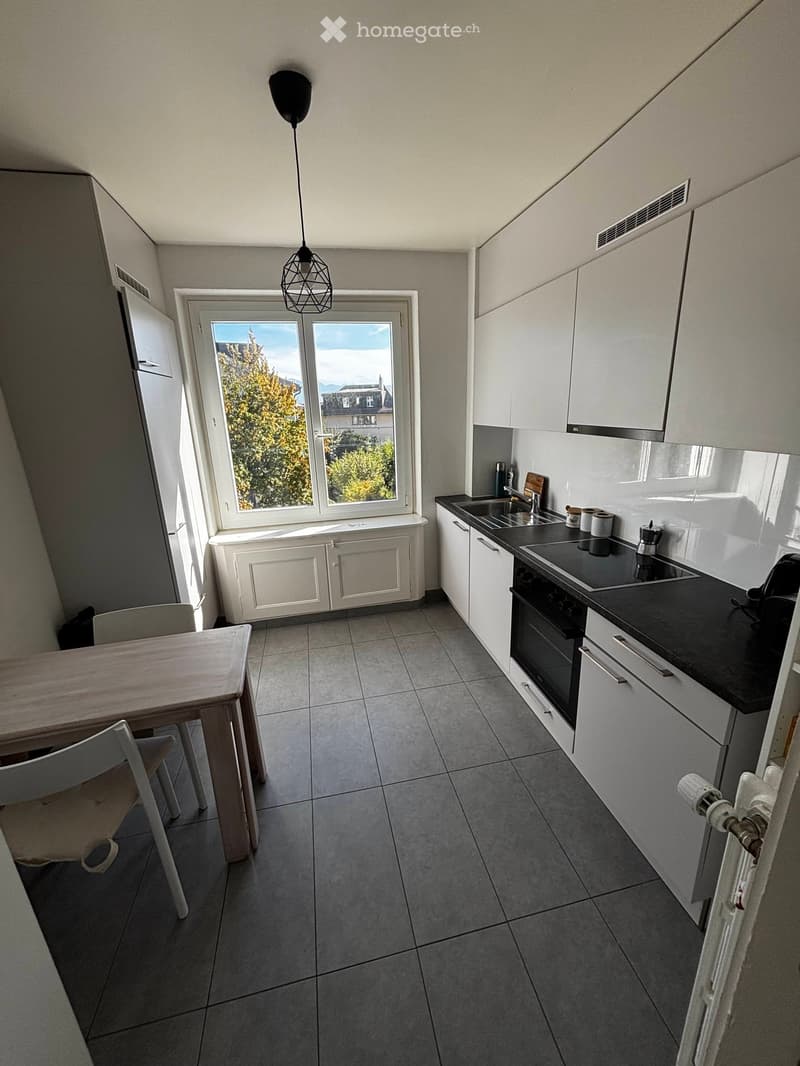 Modern kitchen with white cabinets, black countertops, a built-in oven, sink, and a small dining area with a table and chairs. The room has large windows offering a view of trees and buildings outside.