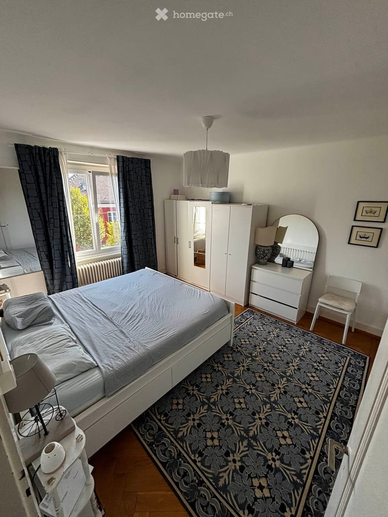 Cozy bedroom with a double bed, white wardrobe, vanity with mirror, nightstand, and a patterned rug.