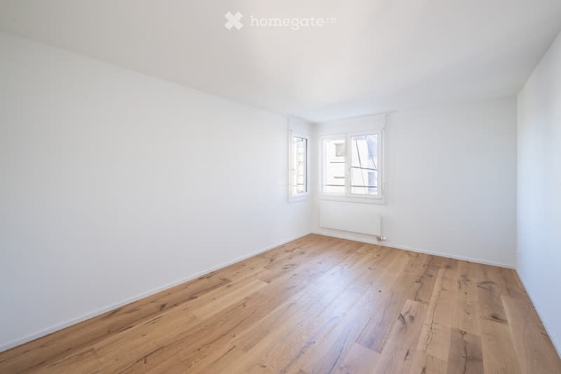 Empty room with white walls, wooden flooring, and two windows with white frames. A radiator is visible under one window.