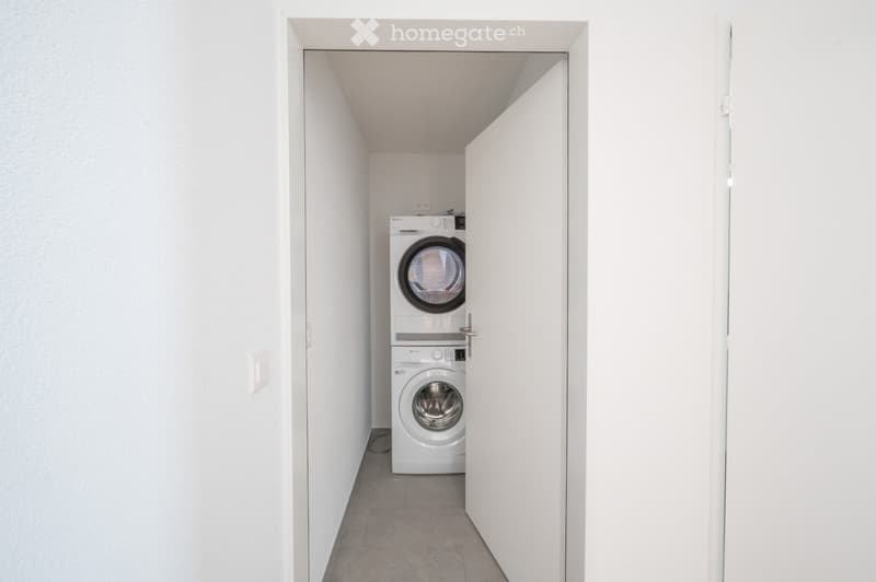 A small, well-lit room with white walls and a tiled floor, featuring a stacked washing machine and dryer unit.
