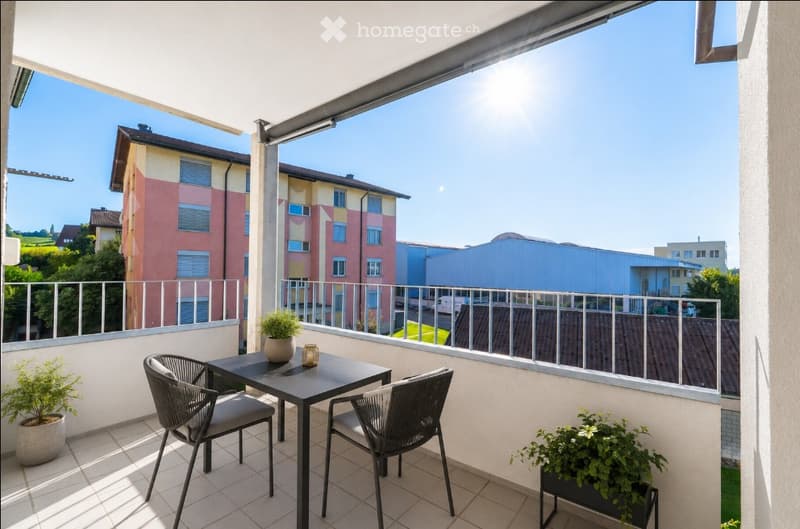 Balcony with a small table and two chairs, potted plants, and a view of surrounding buildings and greenery.
