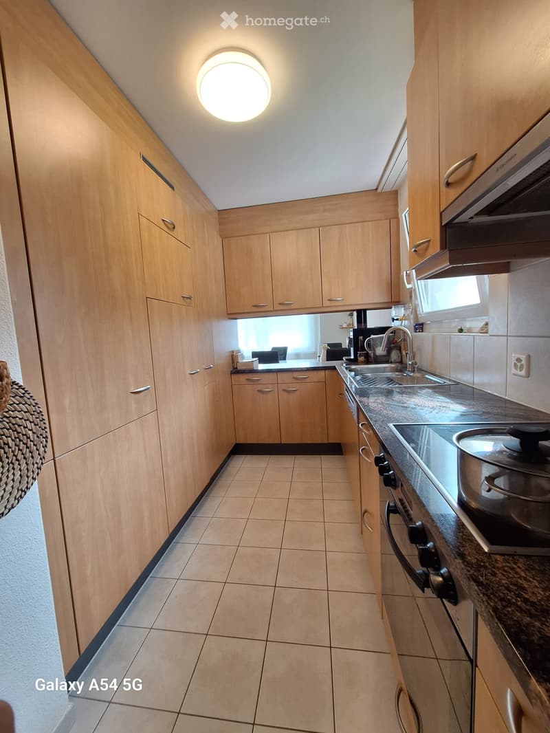 Modern kitchen with light wooden cabinets, granite countertop, stainless steel sink, and built-in appliances including a stove, oven, and microwave. The space features tiled flooring and a circular ceiling light.