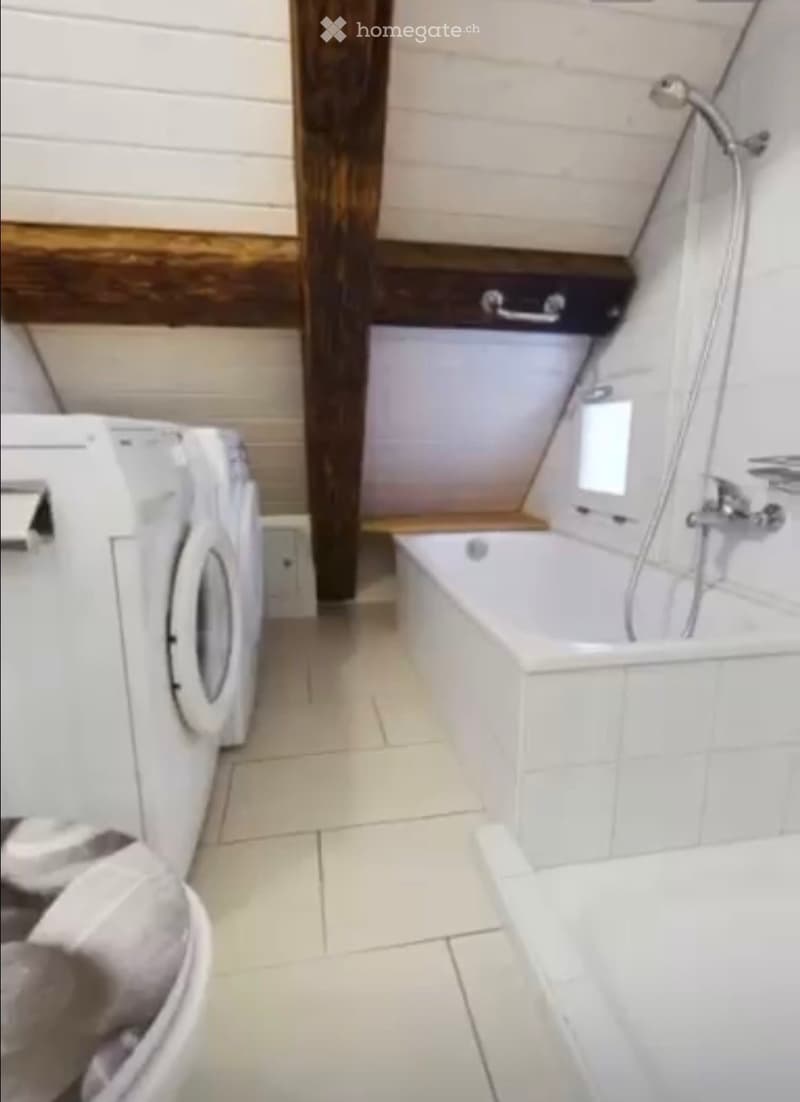 Modern bathroom with a bathtub, shower, and washing machine. The space features white tiled walls and floor, with a wooden accent wall and a built-in shelf.