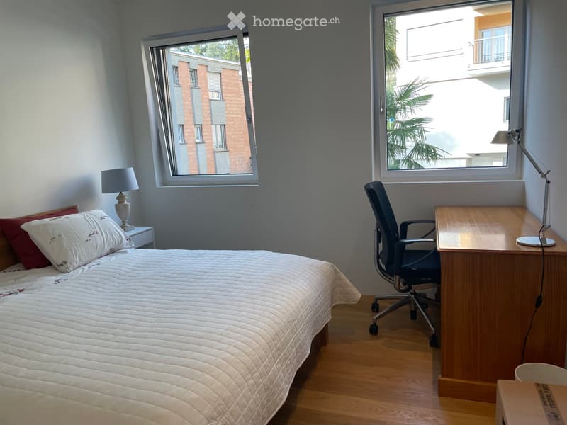 Cozy bedroom with a neatly made bed, a wooden desk with a chair, and a nightstand with a lamp. Large windows offer a view of the building outside.