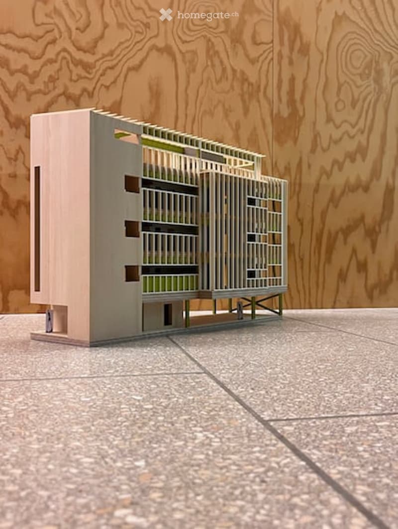 A modern, minimalist building model with a clean geometric design, featuring a combination of light pink and grey materials, set against a wooden backdrop.