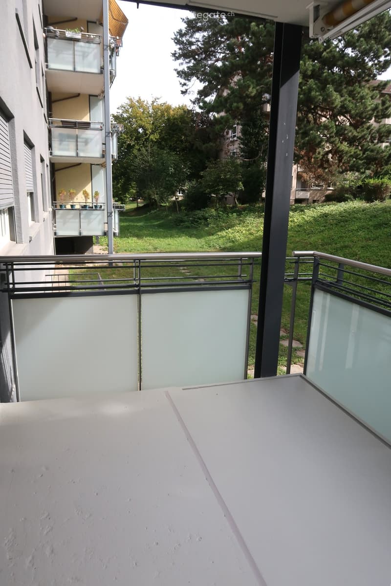 Balcony with modern glass railings, overlooking a green lawn and trees, with adjacent building visible.