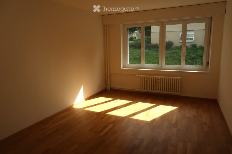 Empty room with wooden flooring, large windows, radiator, and natural light streaming in.