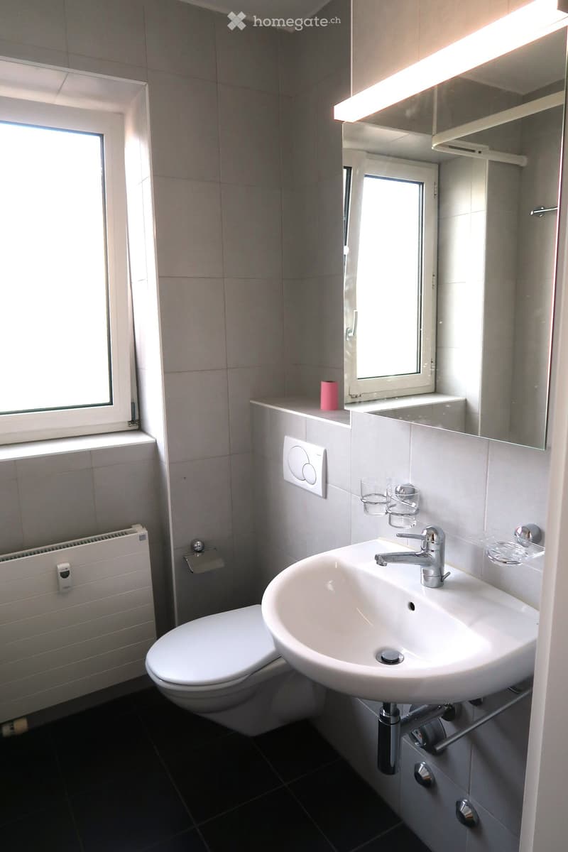 Modern bathroom with white tiles, a toilet, a sink with a chrome faucet, a mirror, and a small window letting in natural light.