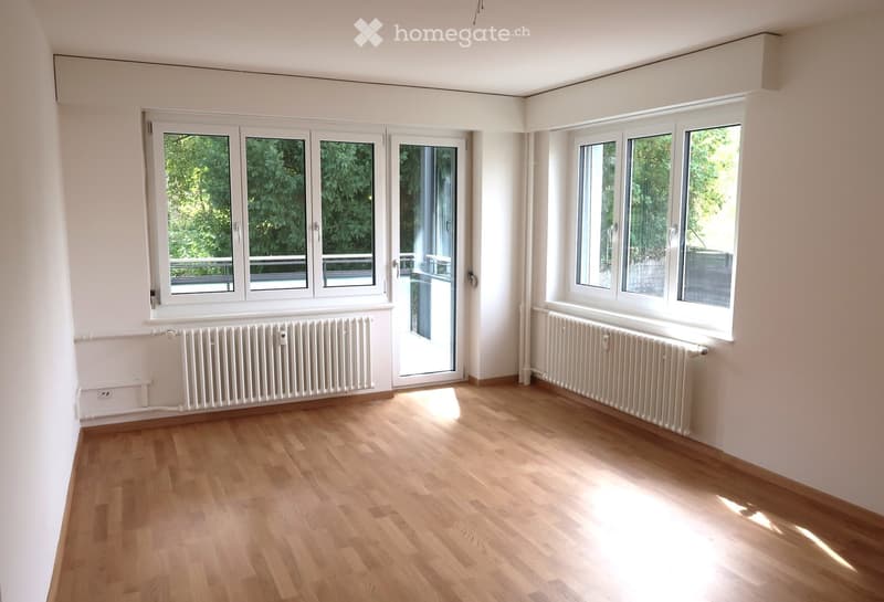 Empty room with wooden flooring, white walls, large windows, radiators, and a sliding glass door leading to a balcony.