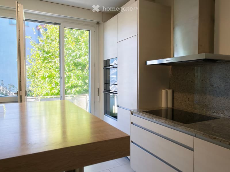 Modern kitchen with white cabinets, a wooden countertop, an oven, and a range hood. The space is bright with large windows and a sliding door leading to a balcony.
