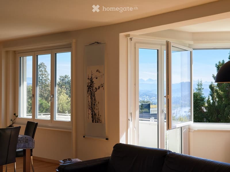 Bright living room with large windows and a view of trees and mountains, featuring a black leather sofa, a wooden dining table with chairs, and a decorative scroll on the wall.