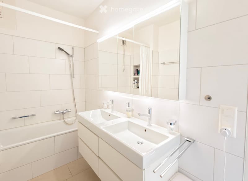Modern bathroom with white tiles, a bathtub, double sink, and a large mirror. Features a shower head and towel rack.