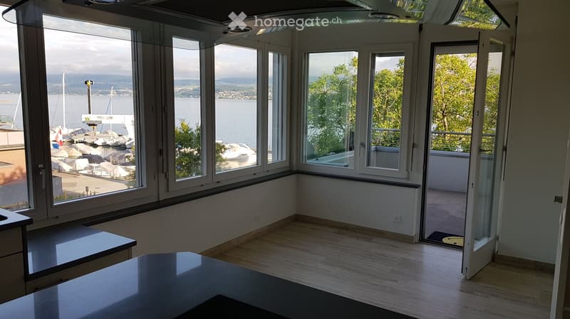 Bright, unfurnished room with large windows offering a scenic view of the water and surrounding area. The room features a wooden floor and a sliding glass door leading to a balcony.