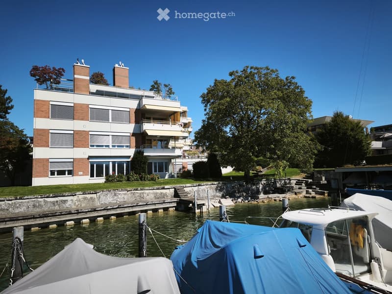 Modern multi-story building with brick and white facade, large windows, balconies, and a waterfront view with docked boats.