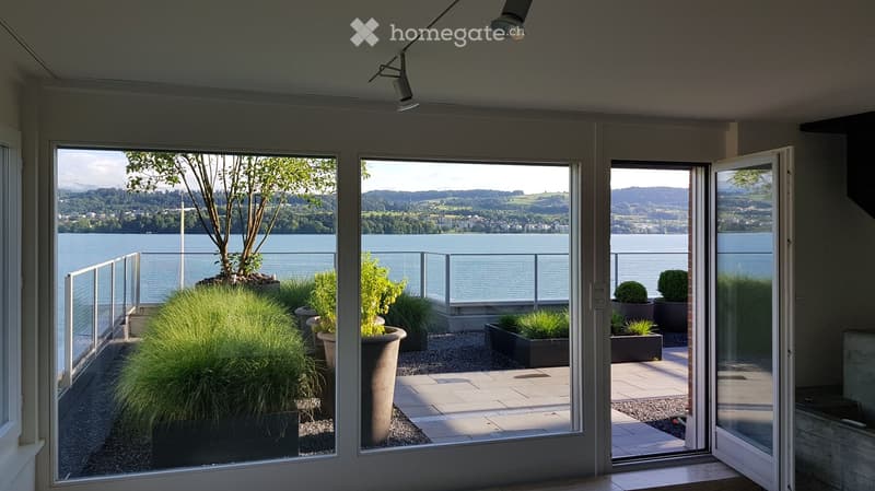 Modern balcony with large glass doors opening to a scenic lake view, featuring potted plants and a paved floor.