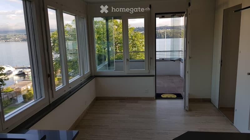 Empty room with large windows overlooking a waterfront, wooden flooring, and an open door leading to a balcony.