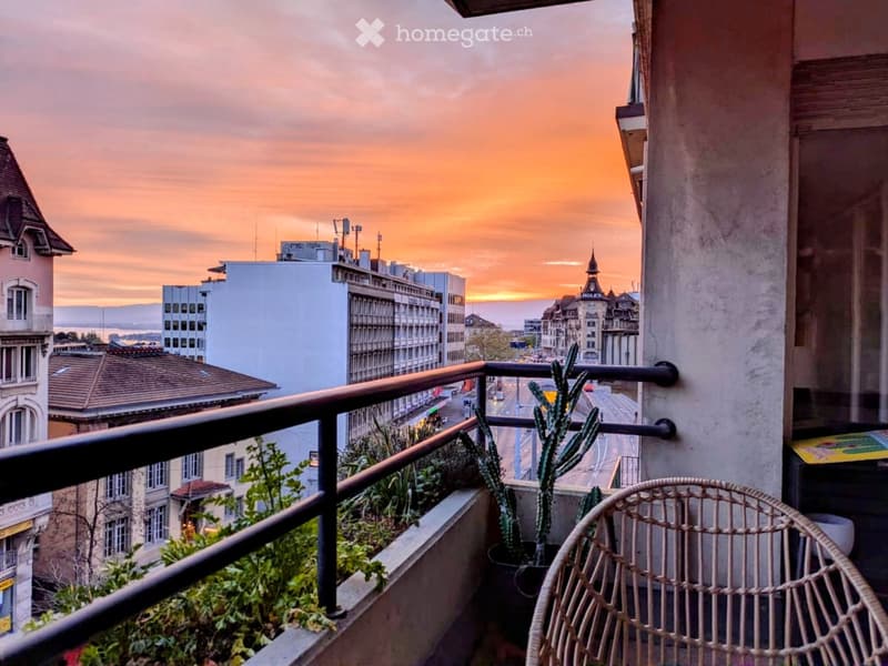 Balcony with a view of the city at sunset, featuring a wicker chair, potted plants, and a metal railing.