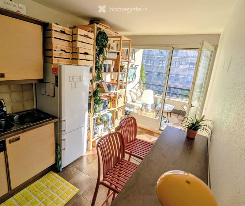 A modern kitchen with wooden cabinets, a refrigerator, a sink, and a dining area with chairs and a table. There is a shelf with jars and a hanging plant, and a view of a balcony with outdoor furniture.