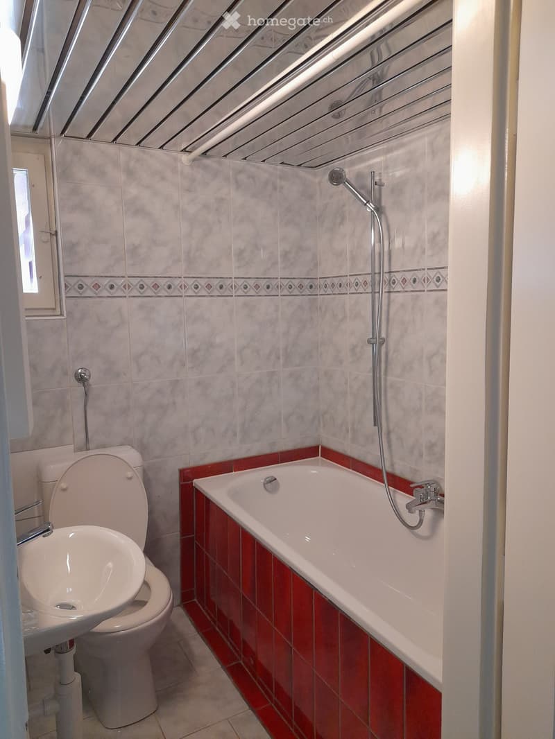 Bathroom with a bathtub, toilet, sink, and a showerhead attached to the wall. The walls are tiled with a decorative border, and the floor is tiled in a light color.