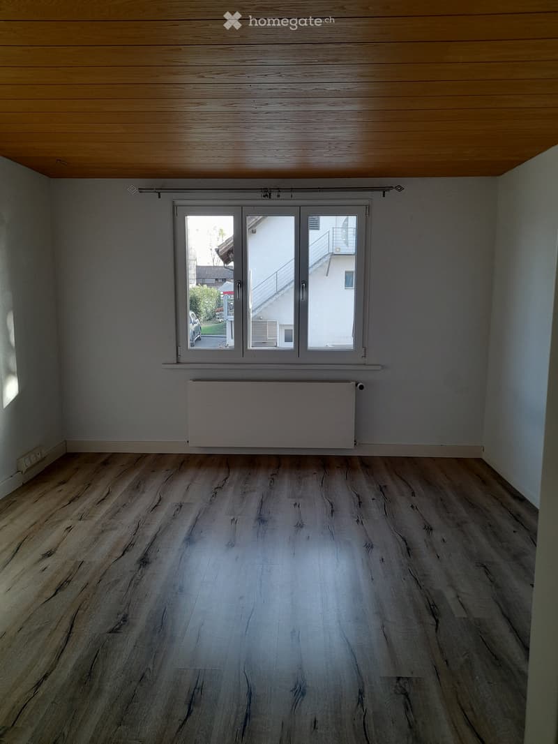 Empty room with wooden flooring, grey walls, large window with sliding doors, radiator, and wooden ceiling beams.